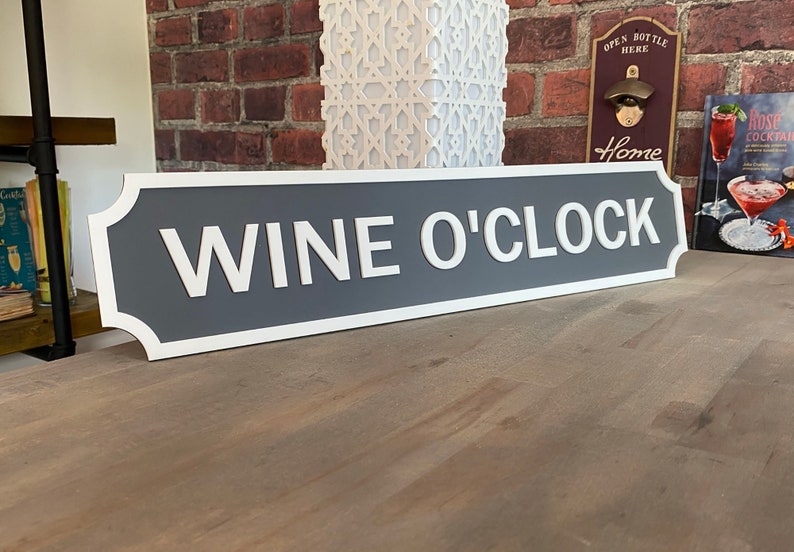 May include: Grey and white sign with rounded edges that reads "Wine O'Clock".