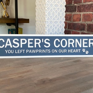May include: A blue and white wooden sign with rounded corners and a white border. The sign reads "CASPER'S CORNER" in white letters and "YOU LEFT PAWPRINTS ON OUR HEART" in white letters with two paw prints.