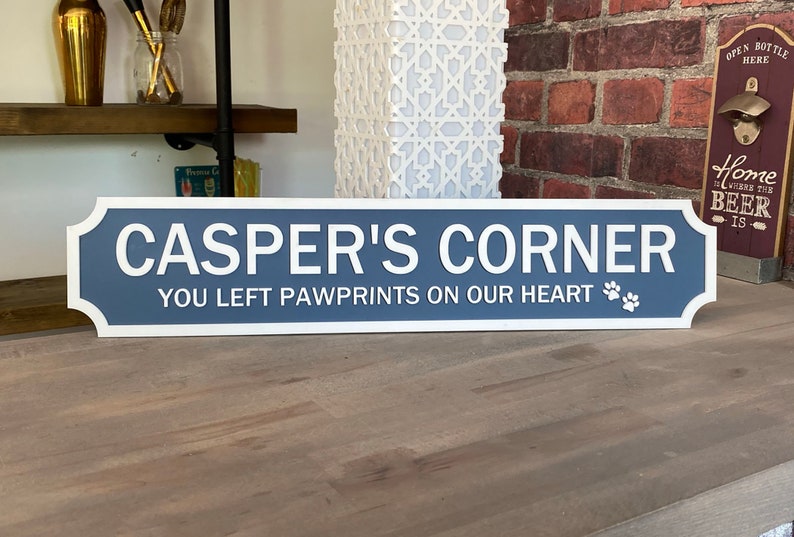 May include: A blue and white wooden sign with rounded corners and a white border. The sign reads "CASPER'S CORNER" in white letters and "YOU LEFT PAWPRINTS ON OUR HEART" in white letters with two paw prints.