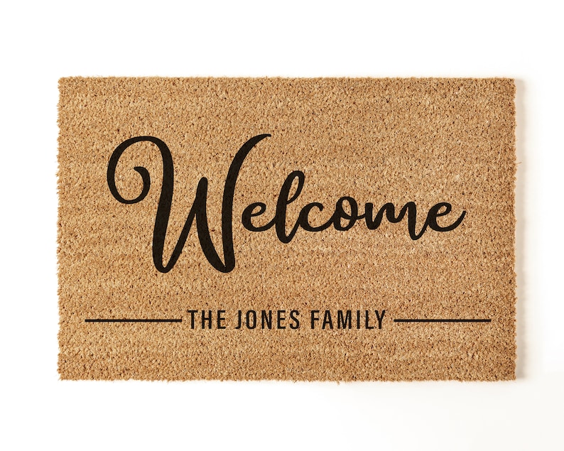 May include: A brown coir doormat with the text "Welcome" in a black cursive font. Below the text is a line with the text "The Jones Family".