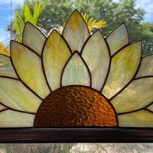 May include: Stained glass sunflower with yellow and gold petals and a brown centre. The glass is framed with dark brown lines. The sunflower is displayed in front of a window with a view of a blue sky and trees.