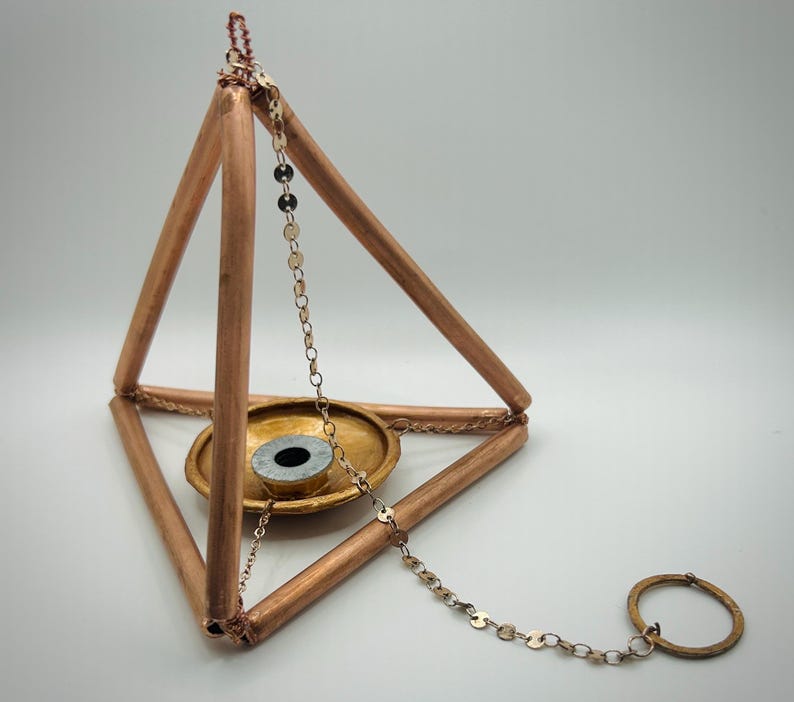 Op de afbeelding: Een koperkleurige piramidevormige wierookhouder met een kleine, ronde, goudkleurige schaal aan de basis. Een zilveren ketting met een ring hangt aan de piramide. De wierookhouder is ontworpen om wierookstokjes of -kegels te bevatten.