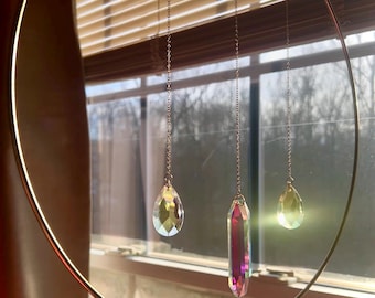 Op de afbeelding: Een goudkleurige metalen hoepel met drie kristallen hangers die aan fijne kettingen hangen. De kristallen, in druppel- en rechthoekige vormen, reflecteren iriserende kleuren. De achtergrond toont een raam met jaloezieën.