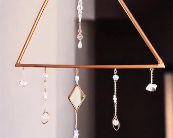 Op de afbeelding: Een geometrische, goudkleurige metalen mobiel met een driehoekig frame. Heldere kristallen en kralen hangen aan fijne kettingen, met een ruitvormige spiegel in het midden. Ontworpen voor interieurdecoratie.