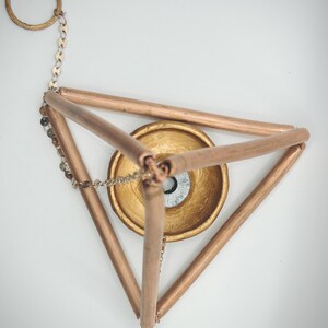 Op de afbeelding: Een geometrische metalen sculptuur in koperkleur. De sculptuur heeft een driehoekig frame met een goudkleurige kom in het midden. Een ketting en ring zijn aan de bovenkant bevestigd. De kom heeft een zilver en zwart ontwerp.