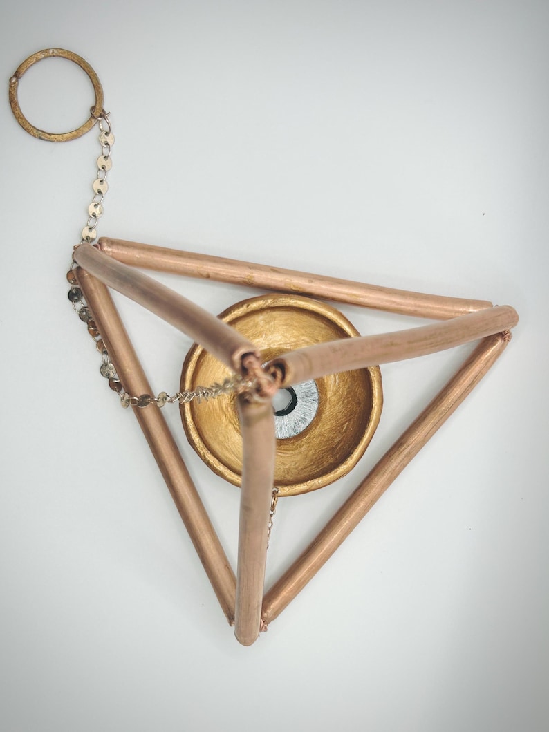 Op de afbeelding: Een geometrische metalen sculptuur in koperkleur. De sculptuur heeft een driehoekig frame met een goudkleurige kom in het midden. Een ketting en ring zijn aan de bovenkant bevestigd. De kom heeft een zilver en zwart ontwerp.