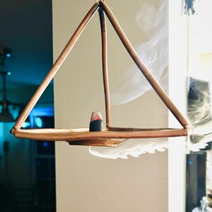 Op de afbeelding: Een koperkleurige wierookhouder in piramidevorm, opgehangen aan kettingen. Een wierookkegel staat op de tray en geeft een wolkje rook af. Het ontwerp is minimalistisch en modern, geschikt voor de interieurdecoratie.