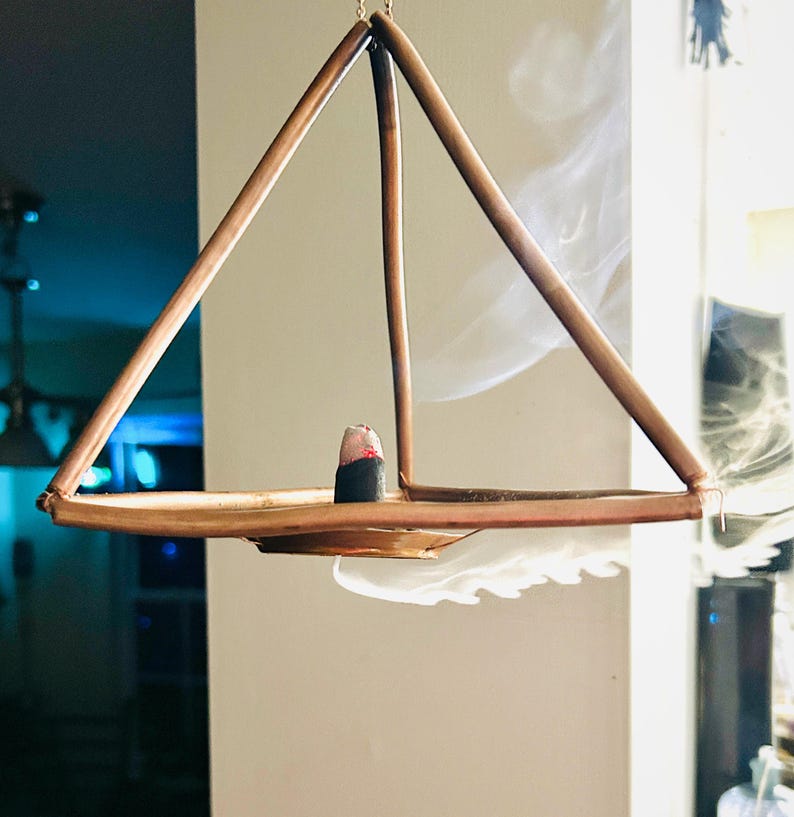 Op de afbeelding: Een koperkleurige wierookhouder in piramidevorm, opgehangen aan kettingen. Een wierookkegel staat op de tray en geeft een wolkje rook af. Het ontwerp is minimalistisch en modern, geschikt voor de interieurdecoratie.