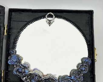 Op de afbeelding: Een zwarte, vierkante sieradendoos met een ronde spiegel. De spiegel is omlijst met een zwarte kanten rand en versierd met blauwe en zilveren rozenversieringen. Een decoratieve zilveren hanger staat bovenaan.