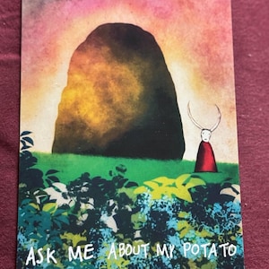 Puede incluir: Una postal con una ilustración de una gran formación rocosa oscura contra un cielo de puesta de sol. Una pequeña figura con una capa roja está cerca. El texto "ASK ME ABOUT MY POTATO" está en la parte inferior.