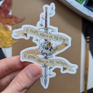 May include: A sticker featuring a sword piercing a skull, with a banner reading "O death, where is the sting?" and "O grave, where is the victory?" The design includes floral elements and is set against a neutral background.