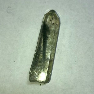 May include: A clear, elongated crystal with a faceted top and a slightly rough, textured surface. The crystal has a smoky, translucent appearance with dark inclusions and a polished, reflective base. The crystal is approximately 3 inches long.