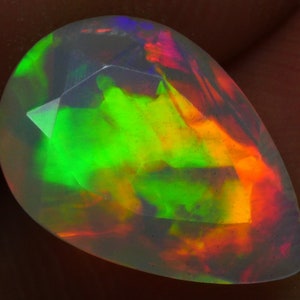 May include: A pear-shaped, faceted opal gemstone. The gem displays a vibrant play-of-color, with flashes of green, yellow, orange, and red. The stone is translucent, allowing light to pass through and create a dazzling effect.