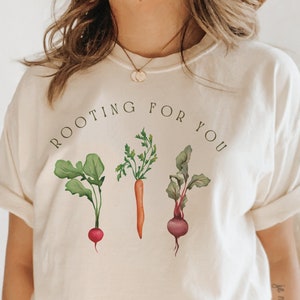 May include: A white t-shirt with the text "ROOTING FOR YOU" in black lettering. The shirt features a graphic of three vegetables: a radish, a carrot, and a beet.