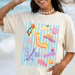 May include: Cream-colored t-shirt featuring a colorful sea slug graphic with blue vertical stripes. The design includes various sea slugs, starfish, and seashells in shades of orange, purple, pink, and yellow. The shirt is a casual, oversized fit.