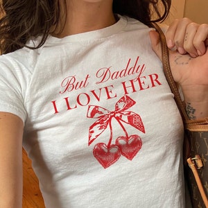 May include: White t-shirt with a red graphic that says "But Daddy I Love Her" and a red cherry design.