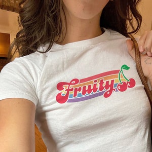 May include: A white t-shirt with a rainbow stripe design and the word "Fruity" in a retro font. Two red cherries are on the right side of the shirt.
