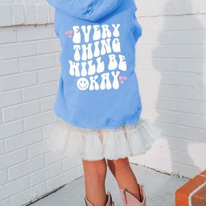 Może przedstawiać: Jas w jasnoniebieskim kolorze z białym nadrukiem "EVERYTHING WILL BE OKAY" z uśmiechniętą buźką i różowymi serduszkami.