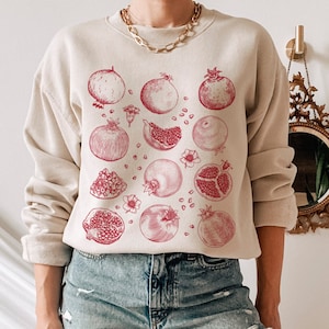 May include: A beige sweatshirt with a red pomegranate and citrus fruit print. The sweatshirt has a crew neck and long sleeves.
