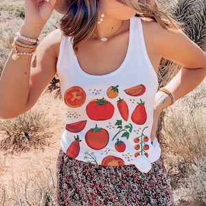 Op de afbeelding: Een vrouw draagt een wit tanktop met een tomaatprint, een bruine hoed en een bloemenrok. Ze staat in een woestijnlandschap.