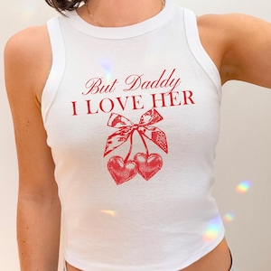 May include: White tank top with a red graphic that says "But Daddy I LOVE HER" with a bow and two heart-shaped cherries. The top is sleeveless and has a round neckline.