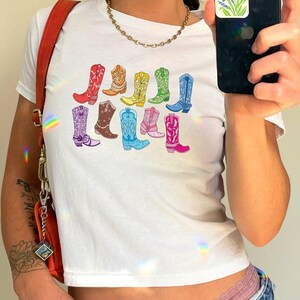 May include: White crop top with a colourful graphic of cowboy boots. The boots are in various colours, including red, orange, yellow, green, blue, and pink. The shirt is paired with blue jeans.