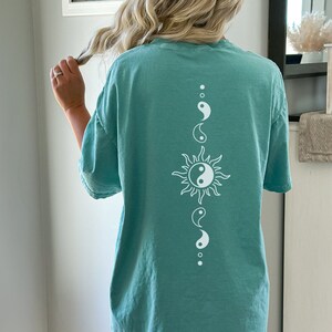 Puede incluir: Una camiseta de gran tama&ntilde;o azul turquesa con un s&iacute;mbolo yin yang blanco y un gr&aacute;fico de sol en la espalda.