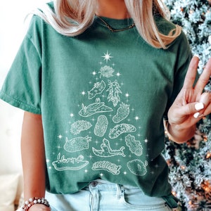 May include: Green t-shirt featuring a white Christmas tree design made of sea slugs and starfish. The shirt is a solid green color and has a relaxed fit. The design is detailed and whimsical, perfect for the holidays.