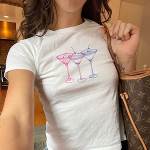 Può includere: Una maglietta bianca con una grafica di tre bicchieri da martini con ciliegie. I bicchieri sono contornati di rosa e blu.