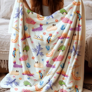 Puede incluir: Una manta crema con un colorido diseño de babosas marinas. El diseño incluye babosas marinas en tonos morados, naranjas, verdes y azules, junto con estrellas de mar y coral. La manta se muestra, revelando el patrón completo.