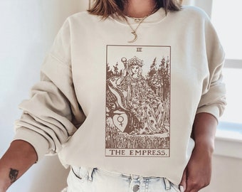 Könnte beinhalten: Ein beiger Pullover mit einem braunen Aufdruck der Tarotkarte der Kaiserin. Die Karte zeigt eine Frau, die auf einem Thron sitzt, eine Krone trägt und ein Zepter hält. Der Text "THE EMPRESS" ist unter der Karte gedruckt.