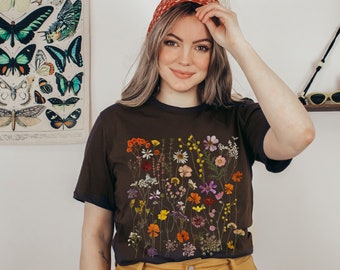 Puede incluir: Una camiseta negra con un estampado floral que presenta una variedad de flores prensadas en tonos de rosa, amarillo, naranja y morado. Las flores están dispuestas en un patrón vertical en la parte delantera de la camiseta.