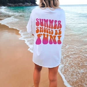 Könnte beinhalten: Weißes T-Shirt mit dem Text "SUMMER SHOULD BE FUN!" in einem Retro-Schriftstil. Der Text wechselt von Pink zu Orange. Das Shirt wird an einem Strand mit dem Meer im Hintergrund getragen.