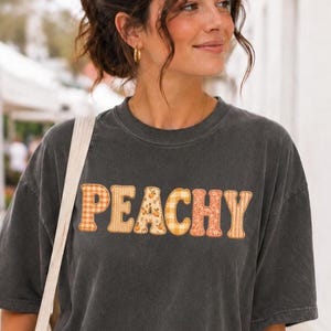 May include: A charcoal gray short-sleeve t-shirt featuring the word PEACHY in large, block-style letters across the chest. Each letter is filled with a different orange and yellow patterned fabric, including gingham, floral, and plaid designs.