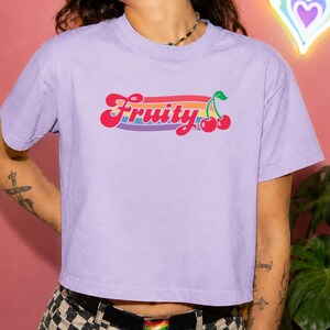 May include: A lilac crop top t-shirt with the word "Fruity" in a retro font, with a rainbow stripe and cherry graphic. The shirt is worn with a black belt and checkered pants. A neon rainbow heart is in the background.