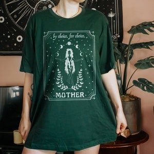 May include: A dark green t-shirt with a white graphic design featuring a woman standing with her arms outstretched. The graphic includes the text "by choice, for choice" and "MOTHER" with a moon and star design.
