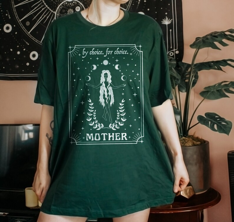 May include: A dark green t-shirt with a white graphic design featuring a woman standing with her arms outstretched. The graphic includes the text "by choice, for choice" and "MOTHER" with a moon and star design.