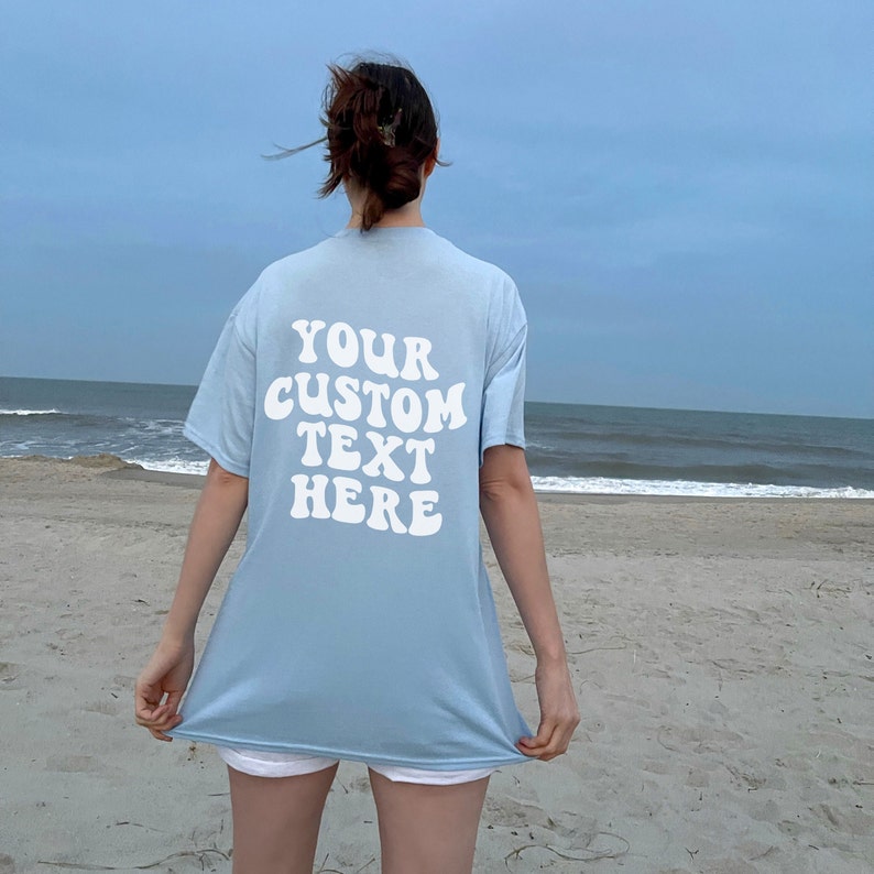 May include: A light blue t-shirt with the text "YOUR CUSTOM TEXT HERE" in white, retro-style lettering on the back. The shirt is being worn by a person standing on a sandy beach.