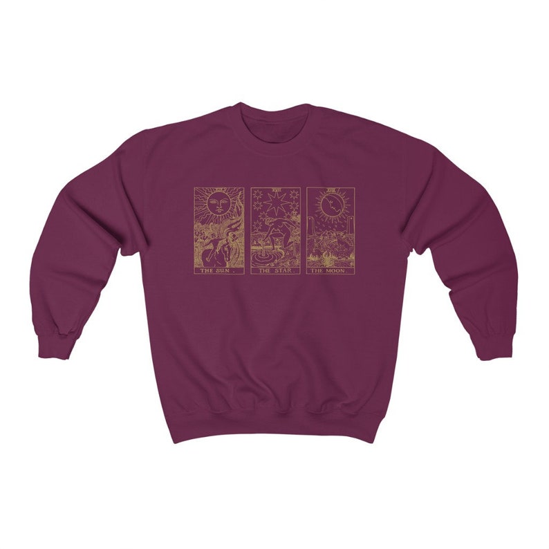 K&ouml;nnte beinhalten: Burgunderfarbenes Sweatshirt mit goldenen Tarotkarten-Designs: Die Sonne, Der Stern und Der Mond. Die Sonnenkarte zeigt eine Sonne mit Gesicht, die Sternenkarte eine Figur, die Wasser gie&szlig;t, und die Mondkarte einen Halbmond.