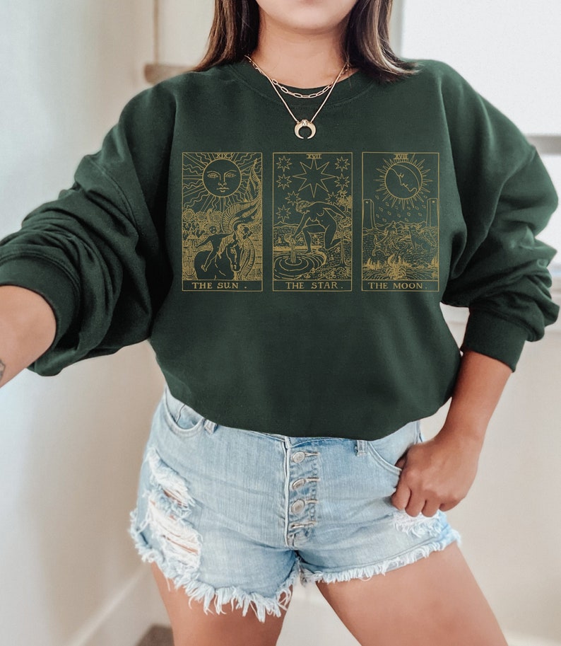 K&ouml;nnte beinhalten: Dunkelgr&uuml;nes Sweatshirt mit goldenen Tarotkarten-Designs: Die Sonne, Der Stern und Der Mond. Die Sonnenkarte zeigt eine Person auf einem Pferd, die Sternenkarte eine Person, die Wasser gie&szlig;t, und die Mondkarte eine Landschaft. Der Text "THE SUN", "THE STAR" und "THE MOON" steht darunter.