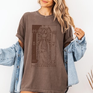 May include: A brown t-shirt with a vintage illustration of the High Priestess tarot card. The card features a woman sitting on a throne with a crown and a veil. The text "THE HIGH PRIESTESS" is printed below the card.