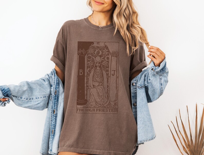May include: A brown t-shirt with a vintage illustration of the High Priestess tarot card. The card features a woman sitting on a throne with a crown and a veil. The text "THE HIGH PRIESTESS" is printed below the card.