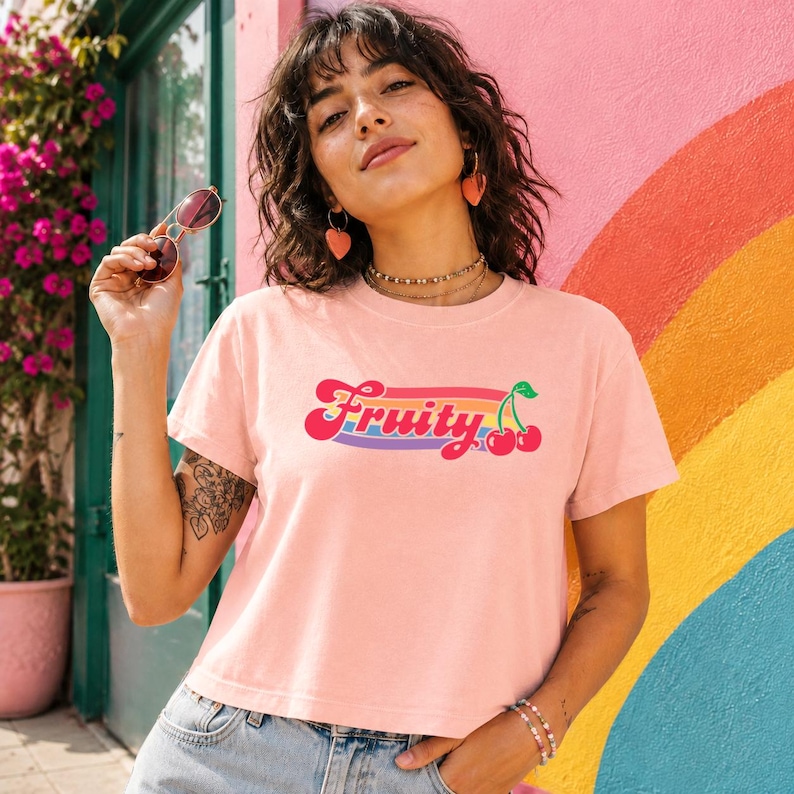 May include: A light pink t-shirt with the word "Fruity" in a retro font, with a rainbow stripe and cherry graphic. The background features a colorful rainbow and a pink wall.
