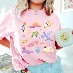 Pu&ograve; includere: Una maglietta rosa con una stampa grafica colorata di varie lumache di mare. La grafica include una variet&agrave; di colori e motivi, tra cui blu, verde, giallo, arancione e viola.