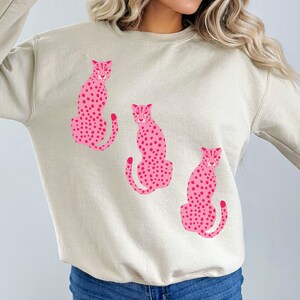 May include: A beige sweatshirt with a pink cheetah print design. The cheetah print is repeated three times on the sweatshirt.