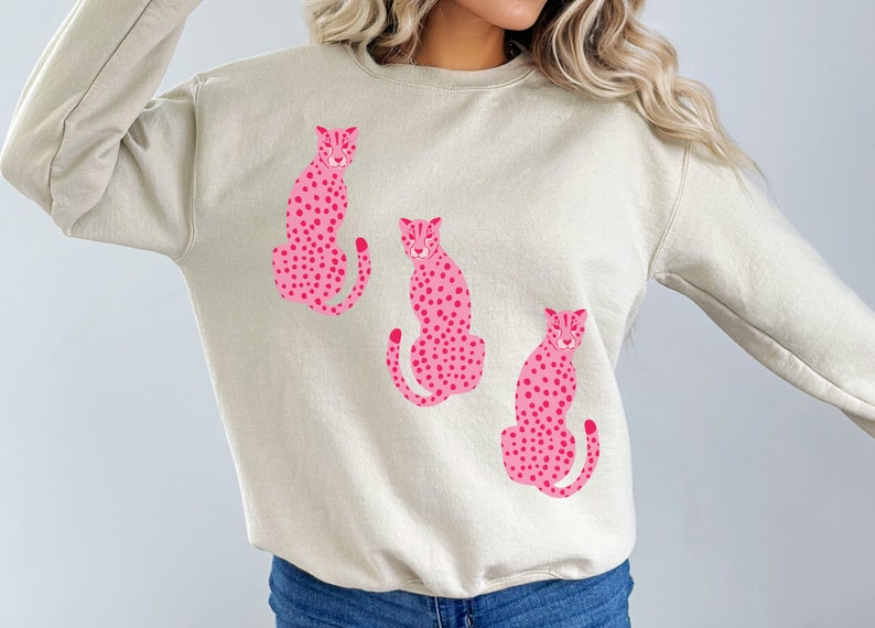 May include: A beige sweatshirt with a pink cheetah print design. The cheetah print is repeated three times on the sweatshirt.
