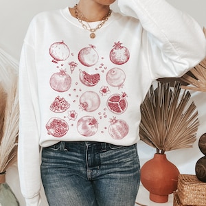 May include: White sweatshirt with a red pomegranate print. The print features multiple pomegranates in various stages of being cut open, showing the seeds inside.