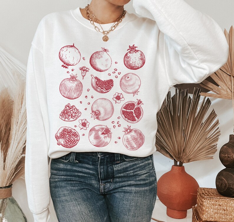 May include: White sweatshirt with a red pomegranate print. The print features multiple pomegranates in various stages of being cut open, showing the seeds inside.