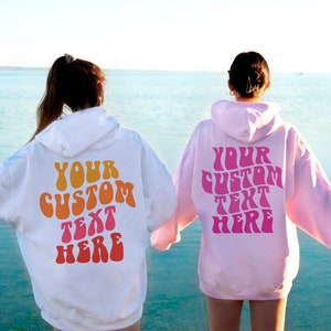 May include: Two people wearing hooded sweatshirts with the text "YOUR CUSTOM TEXT HERE" in a retro font on the back. The sweatshirt on the left is white with orange, yellow, and pink text. The sweatshirt on the right is pink with pink text.