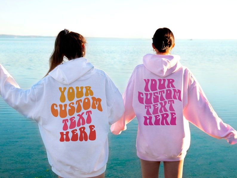 May include: Two people wearing hooded sweatshirts with the text "YOUR CUSTOM TEXT HERE" in a retro font on the back. The sweatshirt on the left is white with orange, yellow, and pink text. The sweatshirt on the right is pink with pink text.